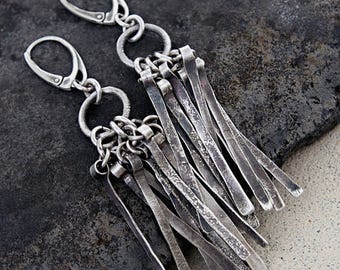 May include: A pair of silver-toned dangle earrings featuring lever-back closures attached to circular rings. Each earring has multiple thin, textured, flat metal strips hanging down in a fringe-like arrangement with a dark, oxidized finish.