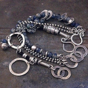 May include: A multi-strand bracelet featuring dark blue beads, silver-toned metal chains, and several circular metal rings. The design includes a cylindrical silver-toned clasp and small metallic spheres interspersed among the beads and chains.