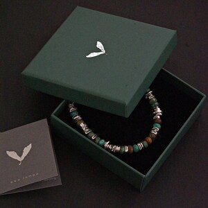 Op de afbeelding: Een kralenarmband met afwisselende turquoise en bruine stenen kralen met zilverkleurige metalen tussenstukjes, gepresenteerd in een donkergroene vierkante doos met een zilveren bladerlogo op het deksel, inclusief een kaartje met de tekst ewa lompe.
