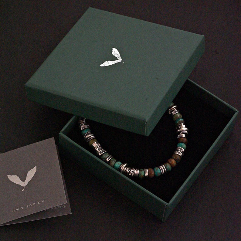 Op de afbeelding: Een kralenarmband met afwisselende turquoise en bruine stenen kralen met zilverkleurige metalen tussenstukjes, gepresenteerd in een donkergroene vierkante doos met een zilveren bladerlogo op het deksel, inclusief een kaartje met de tekst ewa lompe.