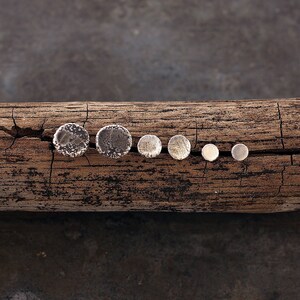 May include: Six silver stud earrings of different sizes are displayed on a weathered wooden log. The earrings vary from large, textured discs to small, smooth circles, creating a rustic and natural look.
