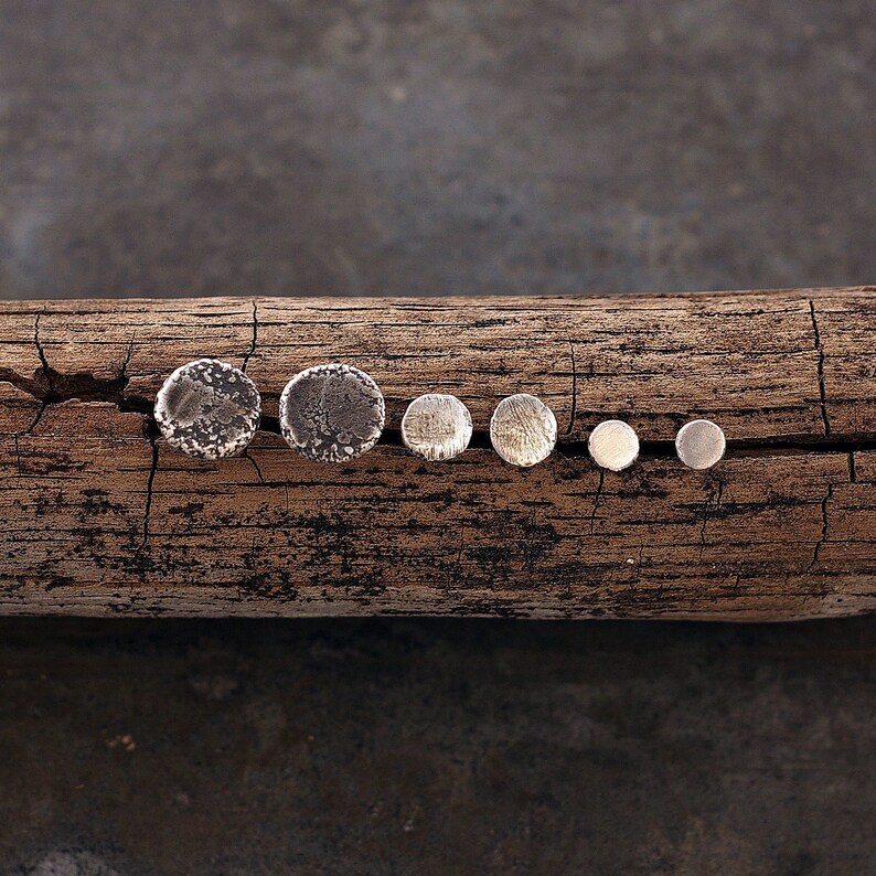 May include: Six silver stud earrings of different sizes are displayed on a weathered wooden log. The earrings vary from large, textured discs to small, smooth circles, creating a rustic and natural look.
