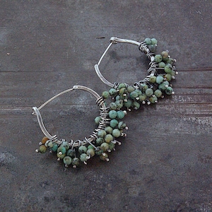 May include: A pair of hoop earrings featuring silver wire frames adorned with numerous small, round, mottled turquoise-colored beads. The beads are densely wrapped and clustered along the lower half of each circular metal hoop structure.