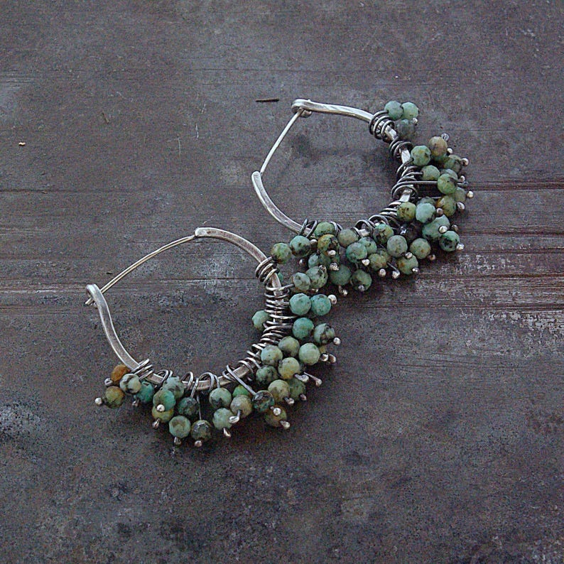 May include: A pair of hoop earrings featuring silver wire frames adorned with numerous small, round, mottled turquoise-colored beads. The beads are densely wrapped and clustered along the lower half of each circular metal hoop structure.