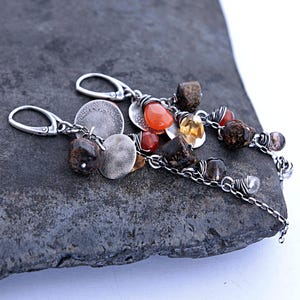 Op de afbeelding: Hangende oorbellen met geoxideerde zilveren kettingen, ronde getextureerde metalen schijven en een mix van gefacetteerde bruine, oranje en heldere edelstenen. De oorbellen zijn voorzien van zilveren klapoorbellen en draadgewikkelde accenten.