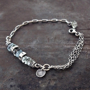 Può includere: Un bracciale a catena in argento con una serie di perline di pietre preziose grezze, sfaccettate e di colore azzurro, intervallate da distanziatori in argento. Il bracciale include una sezione a catena multipla, un moschettone e un ciondolo.