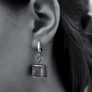 May include: A silver earring featuring a small cube pendant. The earring is attached to a hoop that is connected to the ear. The cube pendant is suspended from the hoop by a small chain. The earring is shown against a blurred background.