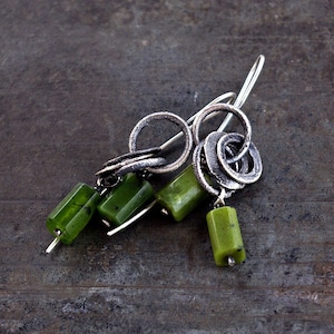 Può includere: Orecchini in argento dal design unico. Ogni orecchino presenta un anello d'argento e un gruppo di tre perline verdi rettangolari. Gli orecchini hanno uno stile moderno e artistico.
