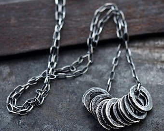 May include: A dark silver-toned necklace featuring a long, textured chain with a cluster of multiple small, circular, washer-like metal rings suspended from the center. The metal has a weathered, oxidized finish with a matte, uneven surface texture.