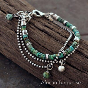 May include: A multi-strand bracelet featuring African turquoise disc beads and small silver-toned metal beads. The design includes a lobster claw clasp, silver-toned spacers, and two dangling charms, one being a round bead and one a smooth metal sphere.