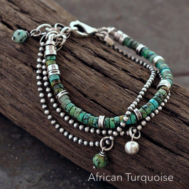 May include: A multi-strand bracelet featuring African turquoise disc beads and small silver-toned metal beads. The design includes a lobster claw clasp, silver-toned spacers, and two dangling charms, one being a round bead and one a smooth metal sphere.
