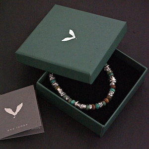 May include: A beaded bracelet featuring alternating turquoise-colored stone beads and metallic silver-toned spacers, housed in a dark green square box with a white leaf logo on the lid. A matching gray card with the text ewa łompe is included.
