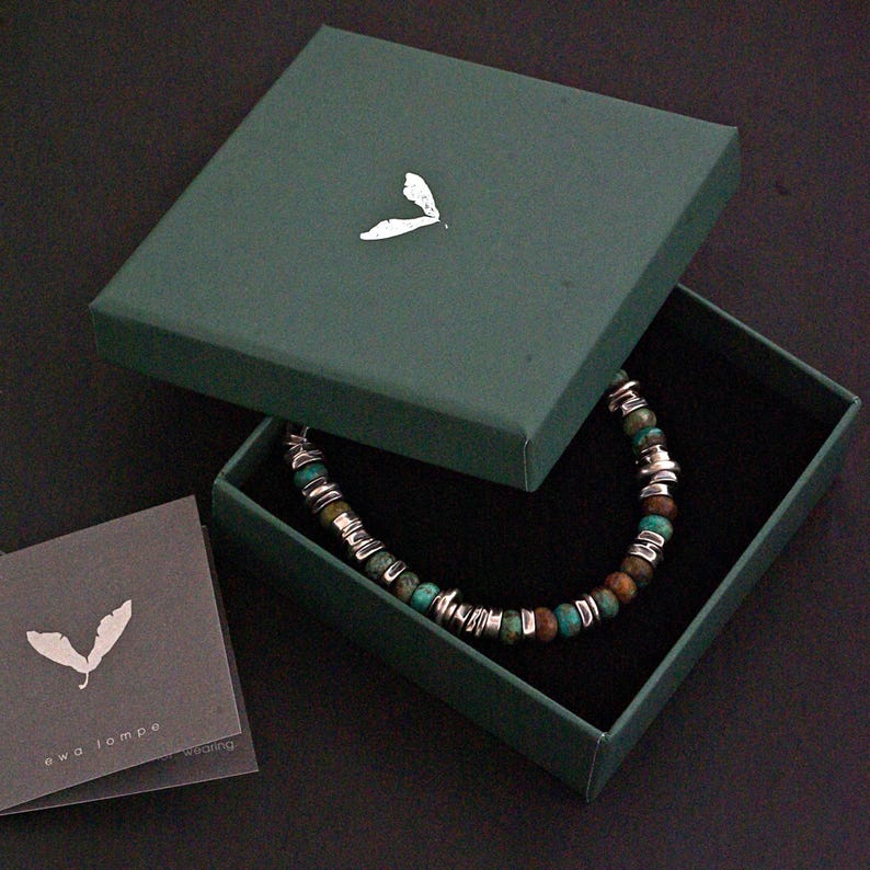 May include: A beaded bracelet featuring alternating turquoise-colored stone beads and metallic silver-toned spacers, housed in a dark green square box with a white leaf logo on the lid. A matching gray card with the text ewa łompe is included.