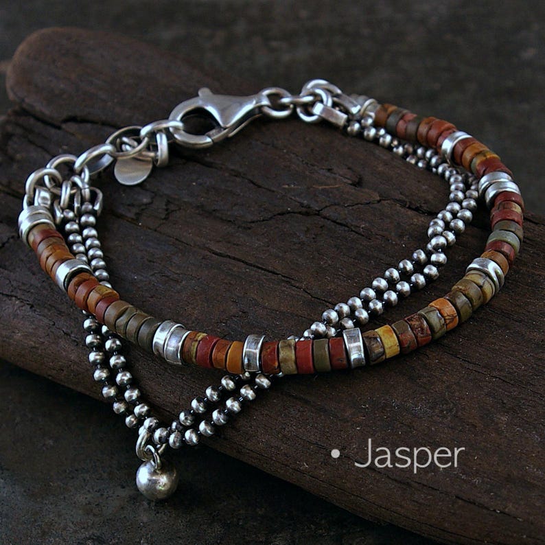 Op de afbeelding: Een armband met meerdere strengen, bestaande uit kleine, ronde, metallic zilveren kralen en platte, schijfvormige jaspis kralen in aardse tinten rood, bruin en beige. De armband bevat zilveren tussenstukjes en een zilveren karabijnsluiting. Tekst: Jasper.