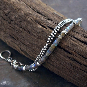 May include: A silver bracelet with two strands. One strand is made of small silver beads, and the other is made of small, colorful, square-shaped beads. The bracelet is displayed on a piece of weathered wood.