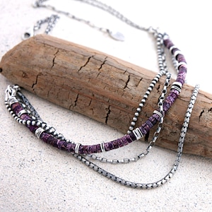 May include: A multi-strand necklace featuring silver-toned metal chains and a strand of small, cylindrical purple stone beads accented with polished silver-toned metal spacers. The necklace includes a delicate silver-toned chain with a small tag.