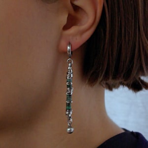 May include: A long drop earring featuring a silver hoop attached to a vertical chain of alternating small dark green cylindrical beads and thin silver spacers, ending with a small silver bead at the bottom, worn on an earlobe.