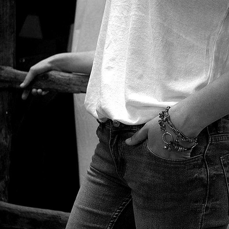 May include: A white sleeveless top paired with dark denim jeans. A multi-strand metallic bracelet with small charms and a circular ring detail is worn on the wrist, partially tucked into the front pocket of the jeans, showcasing a textured design.