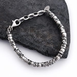 May include: A silver-toned bracelet featuring a series of alternating smooth spherical beads and textured, irregular cylindrical spacers. The piece includes a lobster claw clasp and a small circular tag attached near the chain link closure.