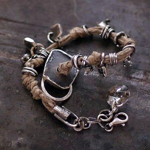 Close-up of a rustic linen bracelet featuring recycled raw silver charms with oxidized, hand-textured surfaces. Each silver piece is melted, shaped, and brushed individually, showing natural imperfections and matte highlights. The linen cord adds softness and earthy contrast, while small crystals introduce gentle, minimal light. A grounded, organic design with visible handmade detail.