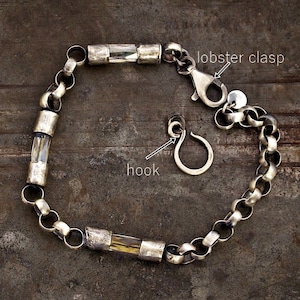 May include: A silver-toned chain bracelet featuring three cylindrical clear crystal segments. The bracelet includes a lobster clasp and a separate hook closure. The metal has a textured, matte finish. The text "lobster clasp" and "hook" point to the components.