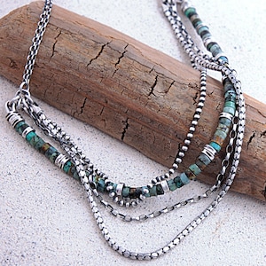 May include: A multi-strand necklace featuring oxidized silver chains and small, cylindrical turquoise beads with brown matrix patterns. The design incorporates smooth silver spacer beads interspersed between the turquoise segments, creating a layered look.