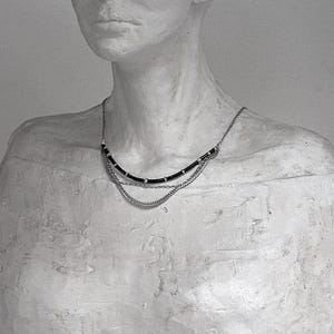 Op de afbeelding: Een meerrijige ketting met een bovenste laag van zwart koord geaccentueerd met kleine zilverkleurige metalen kralen, gecombineerd met twee onderste lagen van fijne zilverkleurige metalen kettingen die over het sleutelbeen vallen.