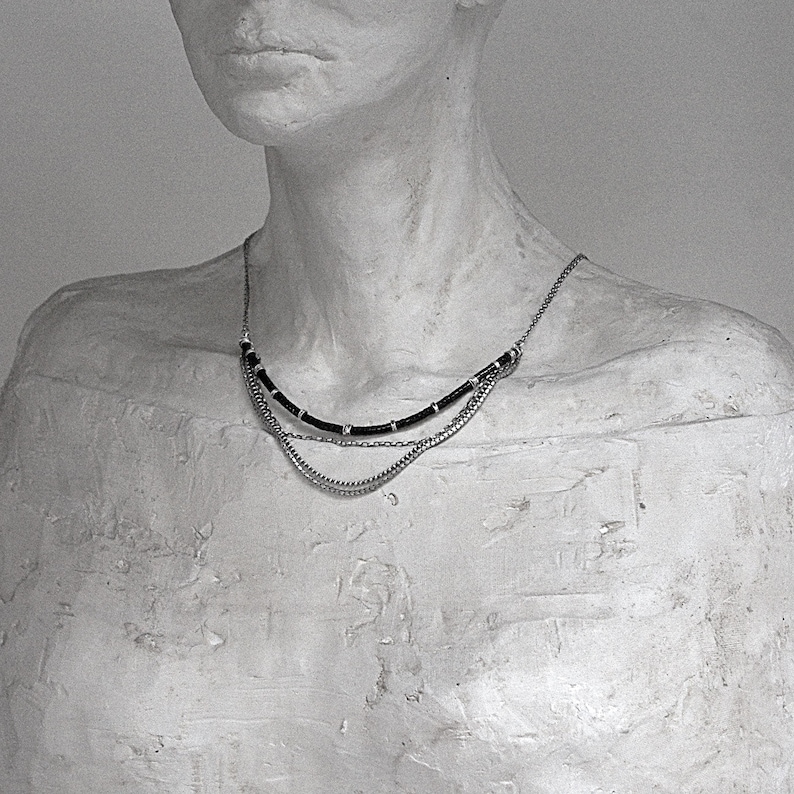 Op de afbeelding: Een meerrijige ketting met een bovenste laag van zwart koord geaccentueerd met kleine zilverkleurige metalen kralen, gecombineerd met twee onderste lagen van fijne zilverkleurige metalen kettingen die over het sleutelbeen vallen.