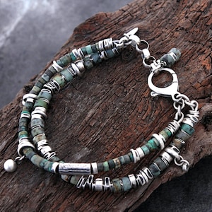 Può includere: Un bracciale a più fili composto da piccole perline cilindriche in pietra dai toni verde e marrone screziato, alternate a distanziatori e anelli in metallo color argento. Il gioiello presenta una chiusura a moschettone argentata e un piccolo ciondolo rotondo.