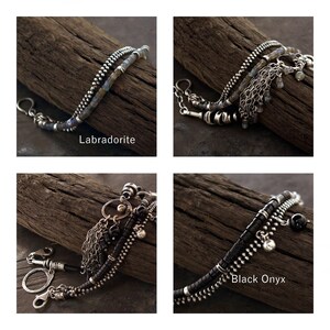 Op de afbeelding: Een armband met meerdere strengen, voorzien van zilverkleurige kettingen en kralen. Er zijn twee versies te zien: een met labradorietstenen en een met zwarte onyxkralen. Beide ontwerpen bevatten metalen bedels, zilverkleurige schakels en een sluiting.