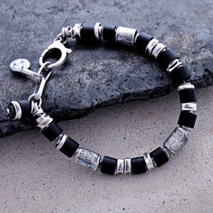 Op de afbeelding: Een armband met zwarte en zilveren kralen. De armband heeft afwisselend zwarte cilindrische kralen en getextureerde zilveren rechthoekige kralen. De sluiting en verbindingsstukken zijn ook zilver.