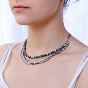 Op de afbeelding: Een meerrijige ketting met &eacute;&eacute;n streng van kleine, groen en zwart gem&ecirc;leerde cilindrische kralen met zilverkleurige tussenstukjes, gelaagd met drie extra dunne, metallic zilverkleurige kralenkettingen.