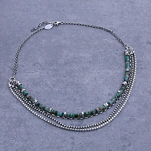 Op de afbeelding: Een meerstrengs ketting bestaande uit zilverkleurige metalen kettingen en &eacute;&eacute;n streng met kleine, ronde, turquoise-kleurige kralen afgewisseld met kleine zilverkleurige metalen tussenstukjes, afgewerkt met een karabijnsluiting.