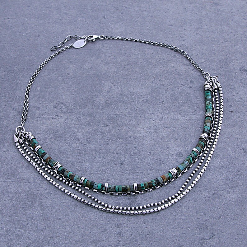 Op de afbeelding: Een meerstrengs ketting bestaande uit zilverkleurige metalen kettingen en &eacute;&eacute;n streng met kleine, ronde, turquoise-kleurige kralen afgewisseld met kleine zilverkleurige metalen tussenstukjes, afgewerkt met een karabijnsluiting.