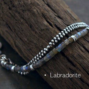Op de afbeelding: Een armband met meerdere strengen, bestaande uit &eacute;&eacute;n streng met kleine, ronde, metallic zilveren kralen en &eacute;&eacute;n streng met vierkante, iriserende labradoriet kralen afgewisseld met zilveren tussenstukjes. De tekst "Labradorite" staat in het wit op de afbeelding.