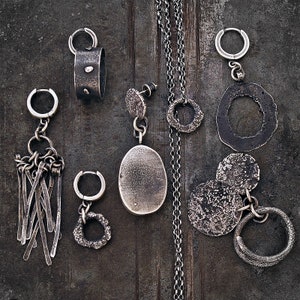 May include: A collection of handcrafted silver jewellery items, including earrings, pendants, and a necklace. The earrings showcase diverse designs, such as textured hoops, dangling bars, and irregular shapes. The necklace features a delicate chain with a textured circular pendant. The jewellery has a rustic, oxidised finish.