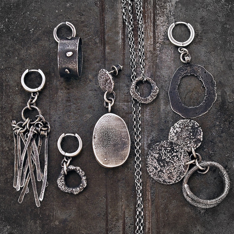 May include: A collection of handcrafted silver jewellery items, including earrings, pendants, and a necklace. The earrings showcase diverse designs, such as textured hoops, dangling bars, and irregular shapes. The necklace features a delicate chain with a textured circular pendant. The jewellery has a rustic, oxidised finish.