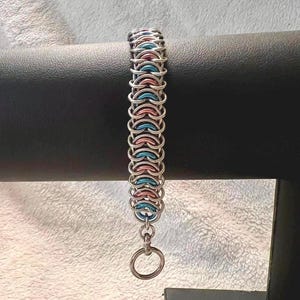 May include: A chainmail bracelet constructed from interlocking metal rings in alternating silver, light blue, and soft pink colours. The bracelet features a repeating pattern of these coloured rings and terminates with a circular metal toggle clasp.