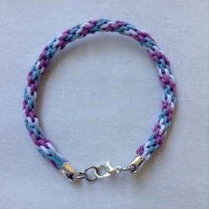 May include: A braided bracelet with a white, light blue, and purple color scheme. The bracelet has a silver clasp.