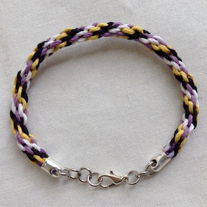 以下が含まれることがあります： 黄色、黒、紫の糸で編まれたブレスレットで、銀色の留め具が付いています。