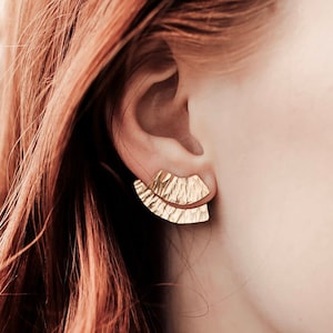May include: A pair of gold-toned earrings with a fan-shaped design. The earrings are made of a textured metal and have a slightly curved shape.
