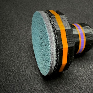 May include: A black and orange sanding disc with a blue and purple foam pad. The disc is designed for use with a rotary tool.