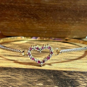 Puede incluir: Una pulsera rígida de tono dorado con un dije central en forma de corazón incrustado con pequeñas piedras rojas y transparentes. Los lados de la pulsera tienen una textura de puntos finos y se unen al corazón mediante pequeñas argollas.