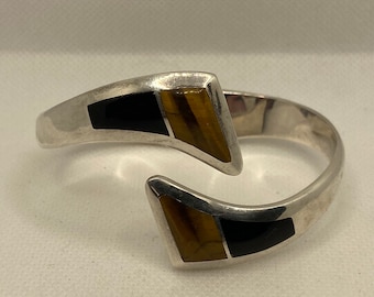 May include: A silver-toned cuff bracelet with geometric accents. The bracelet features black and tiger's eye-colored inlays in a triangular shape. The open design allows for easy wear and a modern aesthetic.