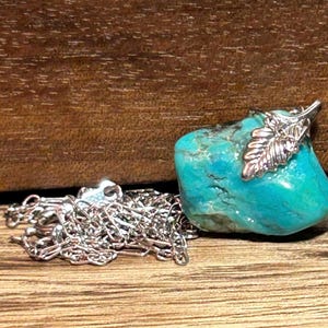 May include: A turquoise stone pendant with a silver leaf accent and a silver chain. The stone is a vibrant blue-green color and has a natural, textured surface. The chain is delicate and shiny.