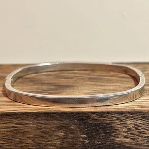 May include: A silver-colored, square-shaped bangle bracelet. The bracelet has a smooth, polished surface and a simple, minimalist design. The bracelet is displayed on a wooden surface.