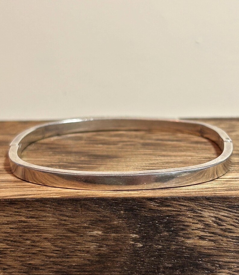 May include: A silver-colored, square-shaped bangle bracelet. The bracelet has a smooth, polished surface and a simple, minimalist design. The bracelet is displayed on a wooden surface.