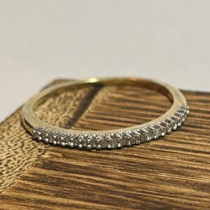 May include: A gold band ring with a row of small, clear gemstones set along the top. The ring is displayed on a wooden surface, showcasing its delicate design and sparkle. The ring is a classic piece of jewelry.