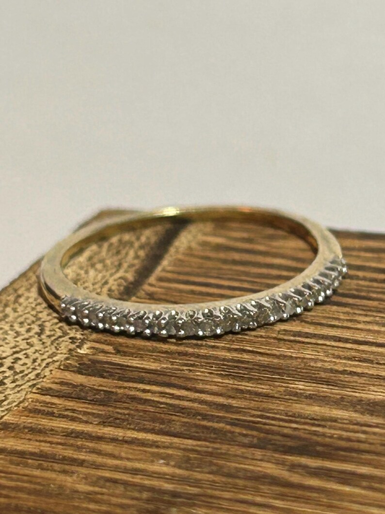 May include: A gold band ring with a row of small, clear gemstones set along the top. The ring is displayed on a wooden surface, showcasing its delicate design and sparkle. The ring is a classic piece of jewelry.