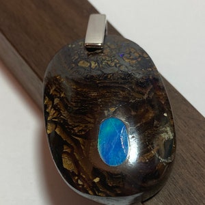 May include: A silver pendant featuring a dark brown stone with a blue opal inlay. The pendant is oval shaped and has a silver bail.